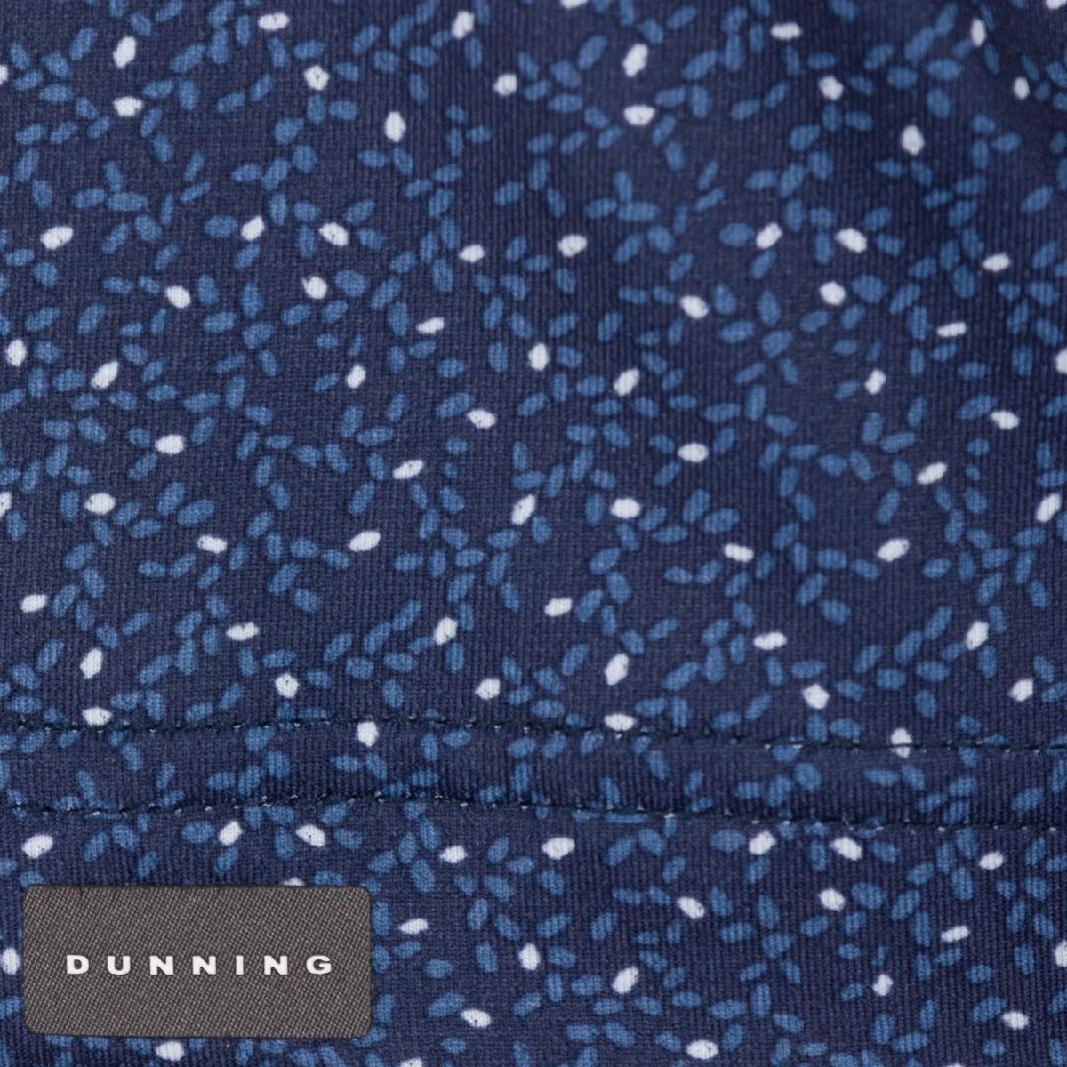 Close-up of a dark Halo blue fabric with a small white and light blue speckled pattern and a stitched seam. A small fabric label at the bottom left reads DUNNING.