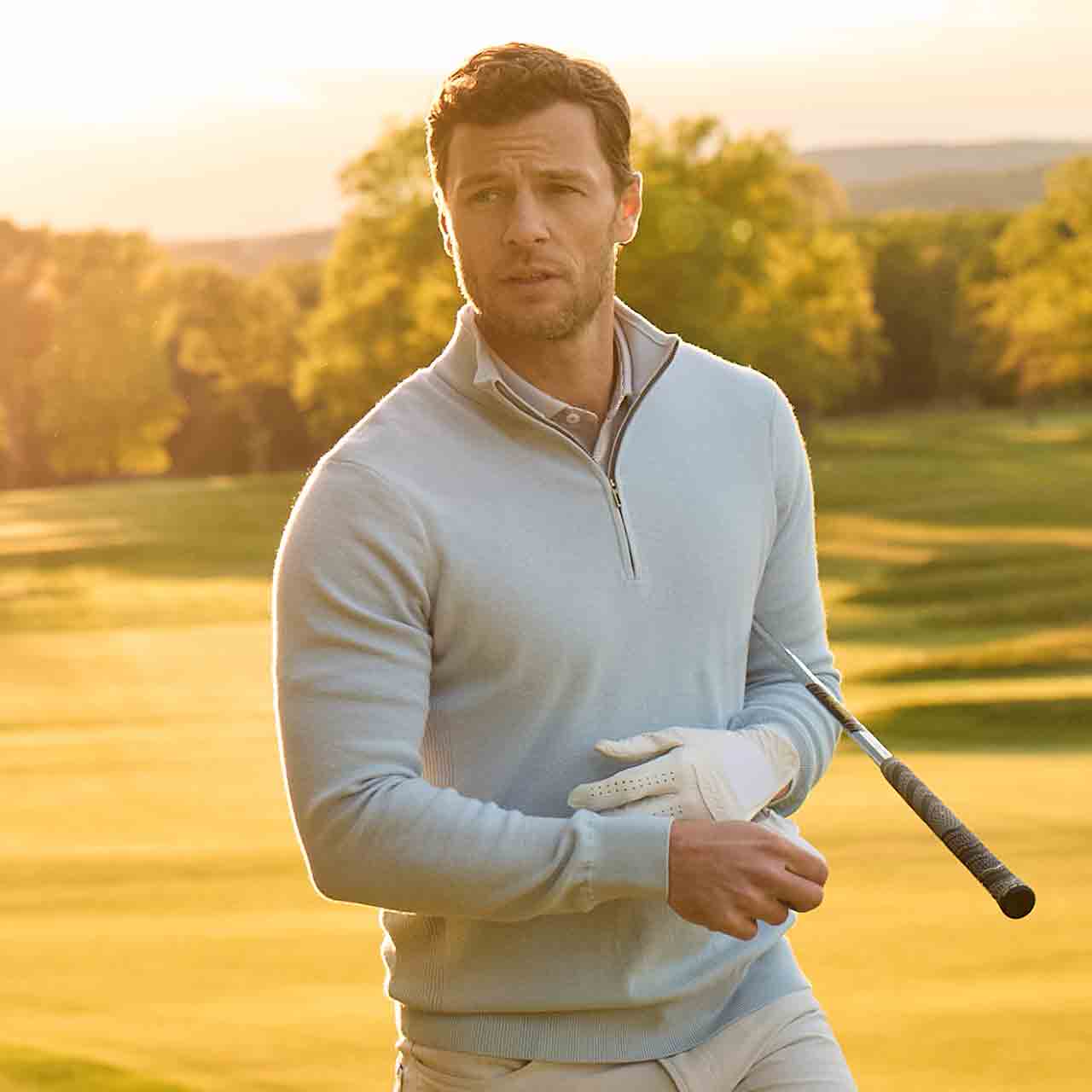 A man wearing a light blue quarter-zip sweater and white golf glove stands on a sunlit golf course holding a golf club.