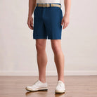 A person wearing Montecarlo Blue shorts with a beige woven belt, standing on a light wood floor against a plain light wall. The person is also wearing white dress shoes and a white top, but the product focus appears to be the shorts.