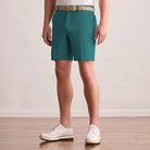 A person wearing Eucalyptus Green golf shorts with a beige woven belt, paired with white wingtip shoes, standing on a light wood floor against a neutral beige wall.