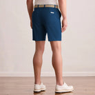 A person shown from the back wearing Montecarlo Blue golf shorts, a beige and blue patterned belt, a white shirt, and white golf shoes on a light wood floor against a neutral beige wall.