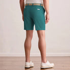 A person shown from the back wearing Eucalyptus Green golf shorts, a tan woven belt, a white shirt, and white golf shoes in a studio setting with a light beige wall and wood floor.
